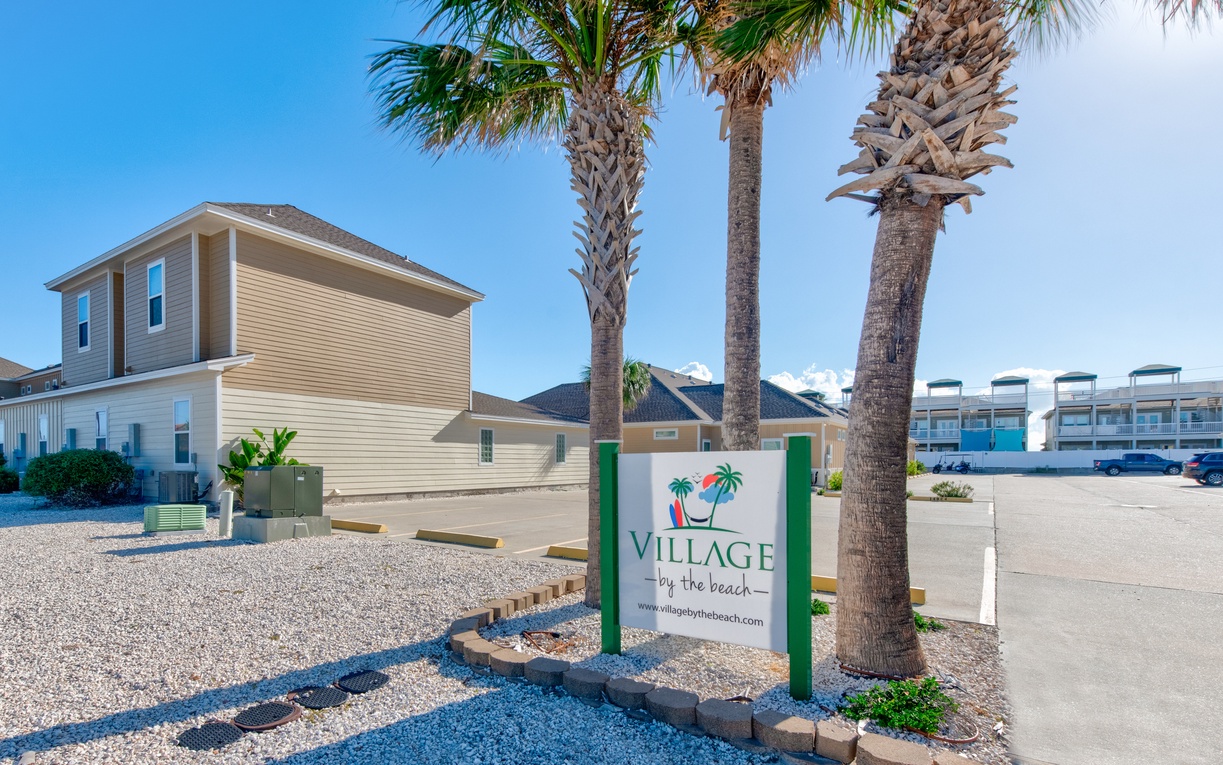 Village by the Beach entrance with coastal palm trees and vacation rental buildings in tropical setting.