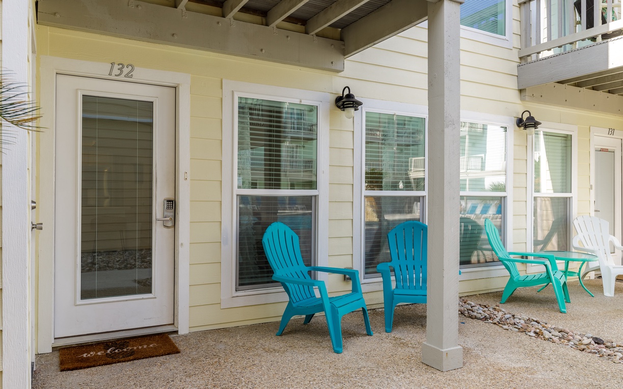 Yellow cottage with covered porch area featuring bright blue Adirondack chairs and white columns, creating a welcoming beach house atmosphere.