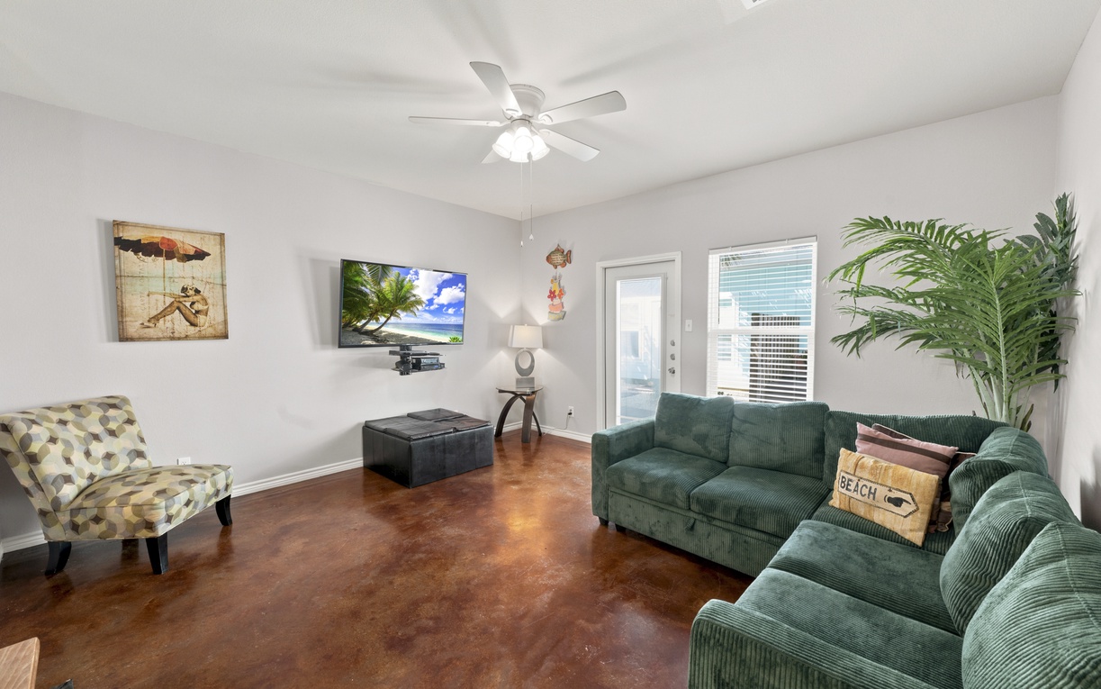 Relax on plush emerald couches as the ceiling fan gently cools your space. This living room offers a cozy retreat with stylish decor, entertainment, and warm, polished floors inviting you to unwind in comfort.
