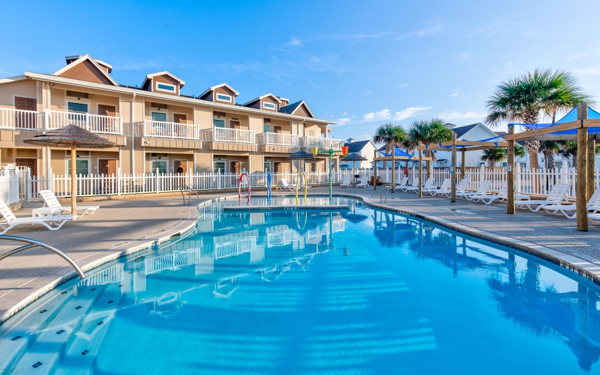 Waterfront resort featuring multi-story accommodations with balconies overlooking a central swimming pool area surrounded by palm trees and lounge chairs.
