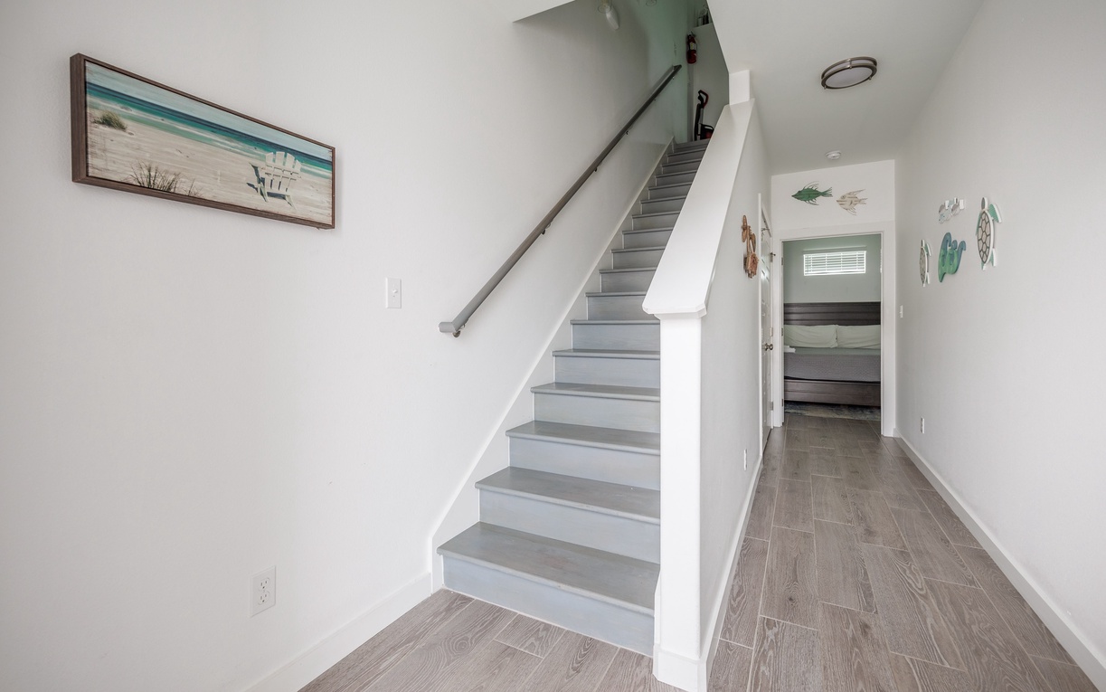 Bright, coastal-themed entryway with elegant stairs leading to your upstairs retreat, featuring beautiful beachside artwork.