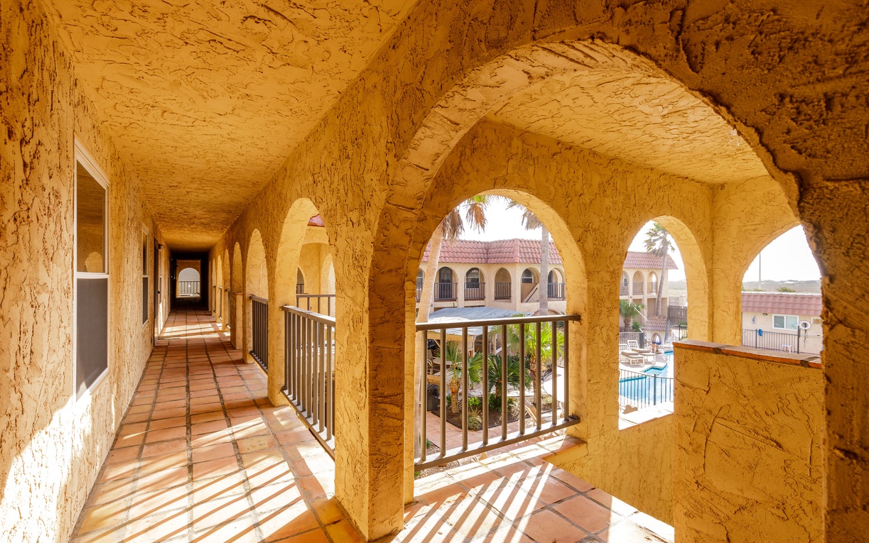 Stone arches frame your pathway to relaxation, where warm terracotta tiles lead you through this sun-dappled corridor toward peaceful courtyards below.