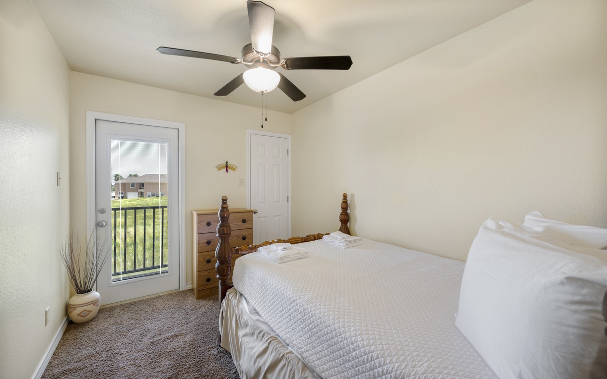 Relax in your cozy bedroom featuring a plush bed, ceiling fan for comfort, and a private balcony door leading to outdoor views. Unwind in this peaceful retreat.