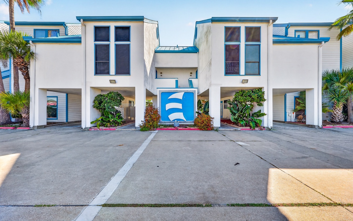 Modern vacation property with convenient covered parking, tropical landscaping, and easy access. The building features white exterior walls with blue trim and ground-floor garage spaces.