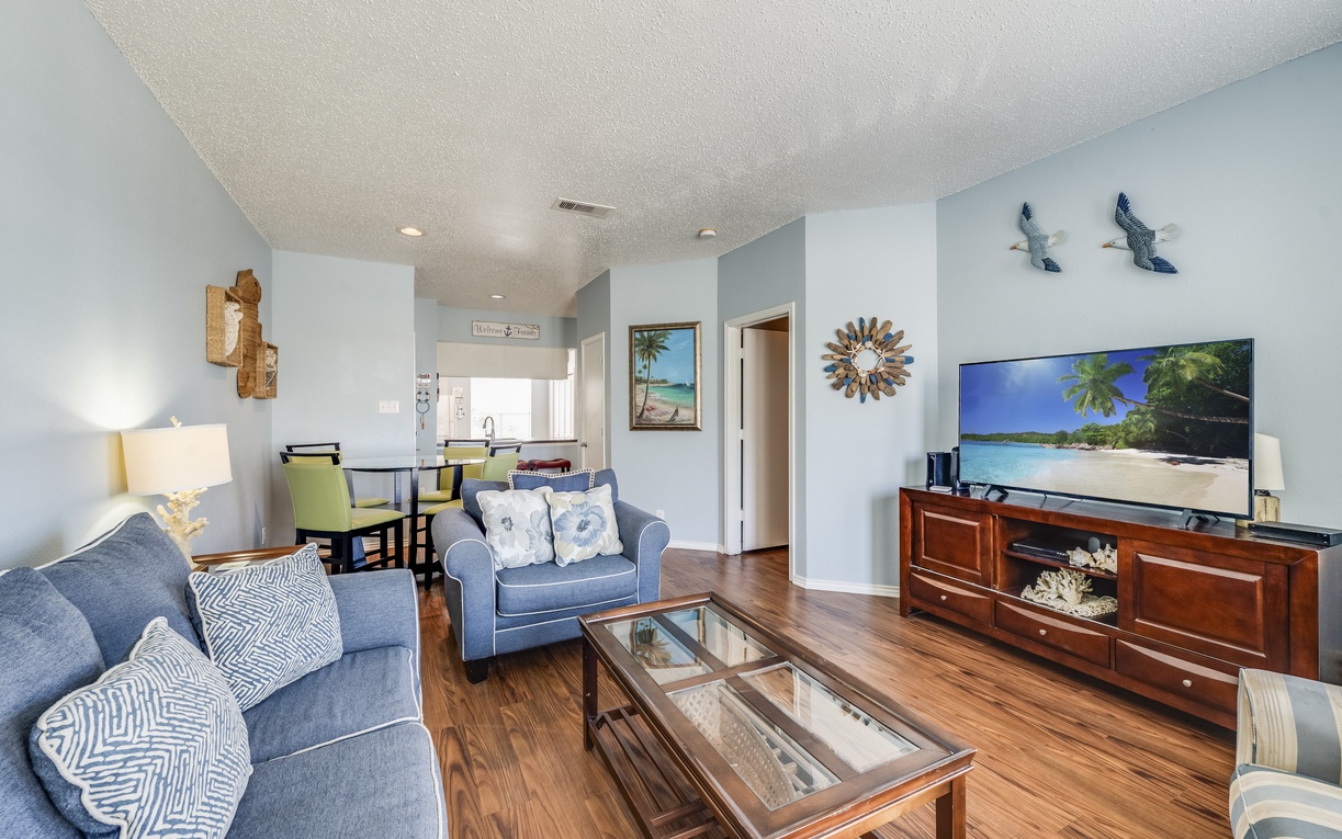 Relax in your spacious living room with comfortable blue seating and tropical beach views on the large TV.