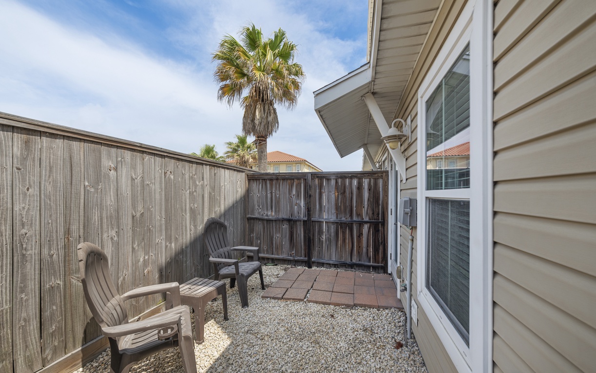 Step out to your private patio featuring comfortable outdoor seating on gravel flooring, surrounded by wooden privacy fencing with palm trees overhead.