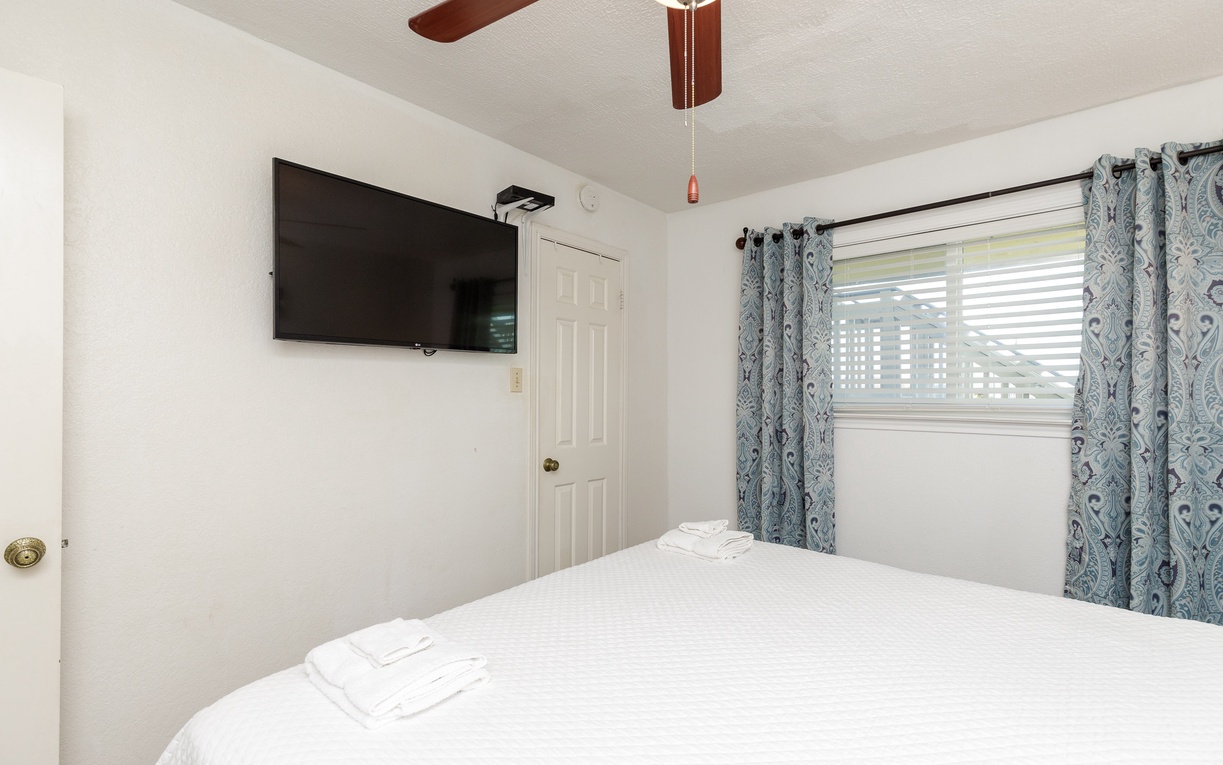 Settle into your cozy bedroom with crisp white linens, a wall-mounted TV for relaxation, and peaceful views through patterned curtains.