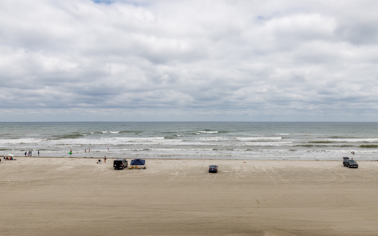 Wide sandy beach stretches along the coastline with gentle waves rolling in under a cloudy sky.