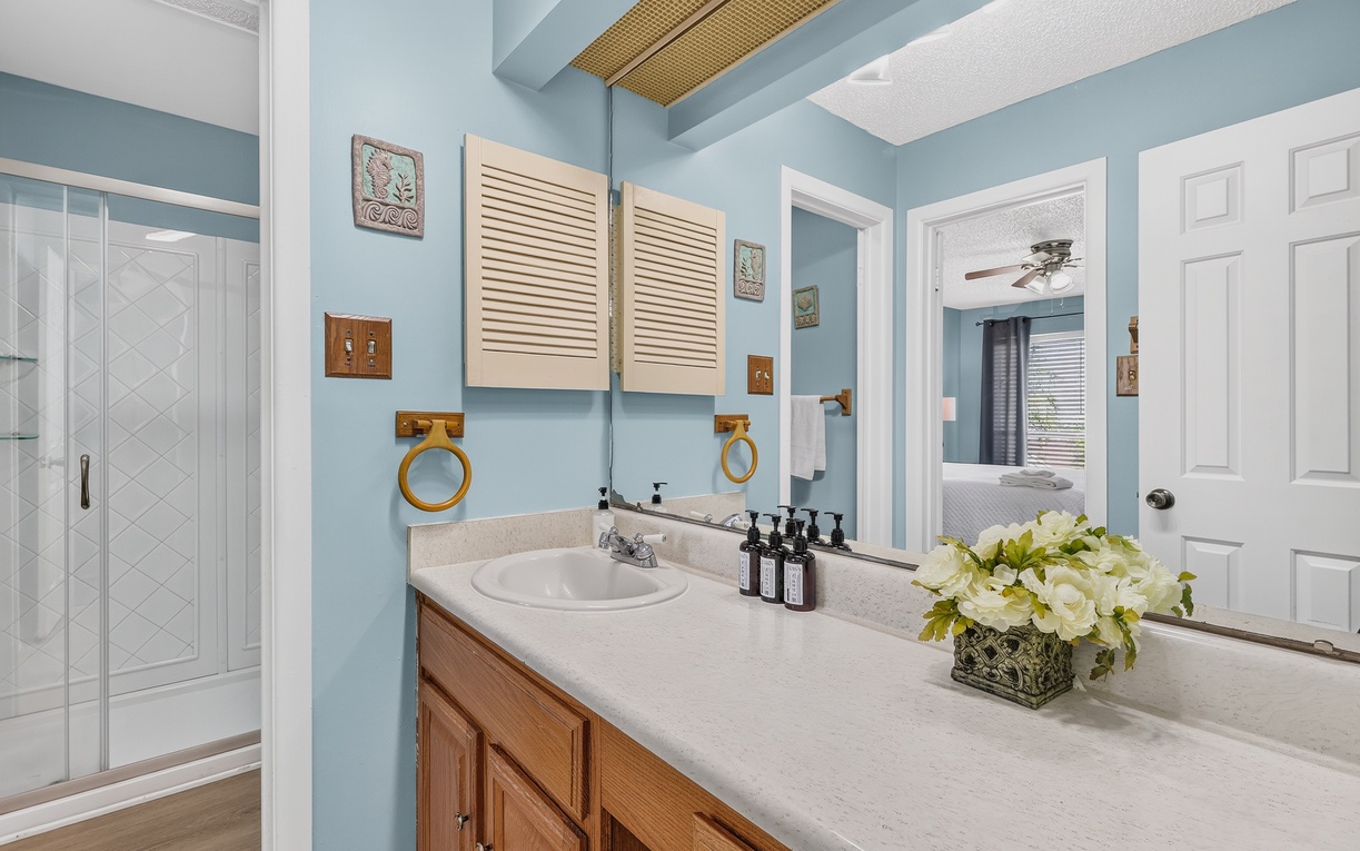 Step into your serene bathroom oasis with calming coastal-blue walls and a spacious vanity. Fresh flowers and luxurious amenities await, while natural light from the adjoining bedroom creates a bright and welcoming retreat.