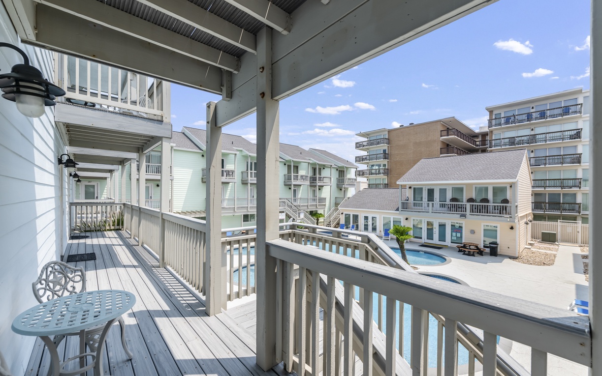 A covered balcony overlooks a serene pool courtyard, framed by coastal-style residences and a backdrop of mid-rise buildings under a bright blue sky.