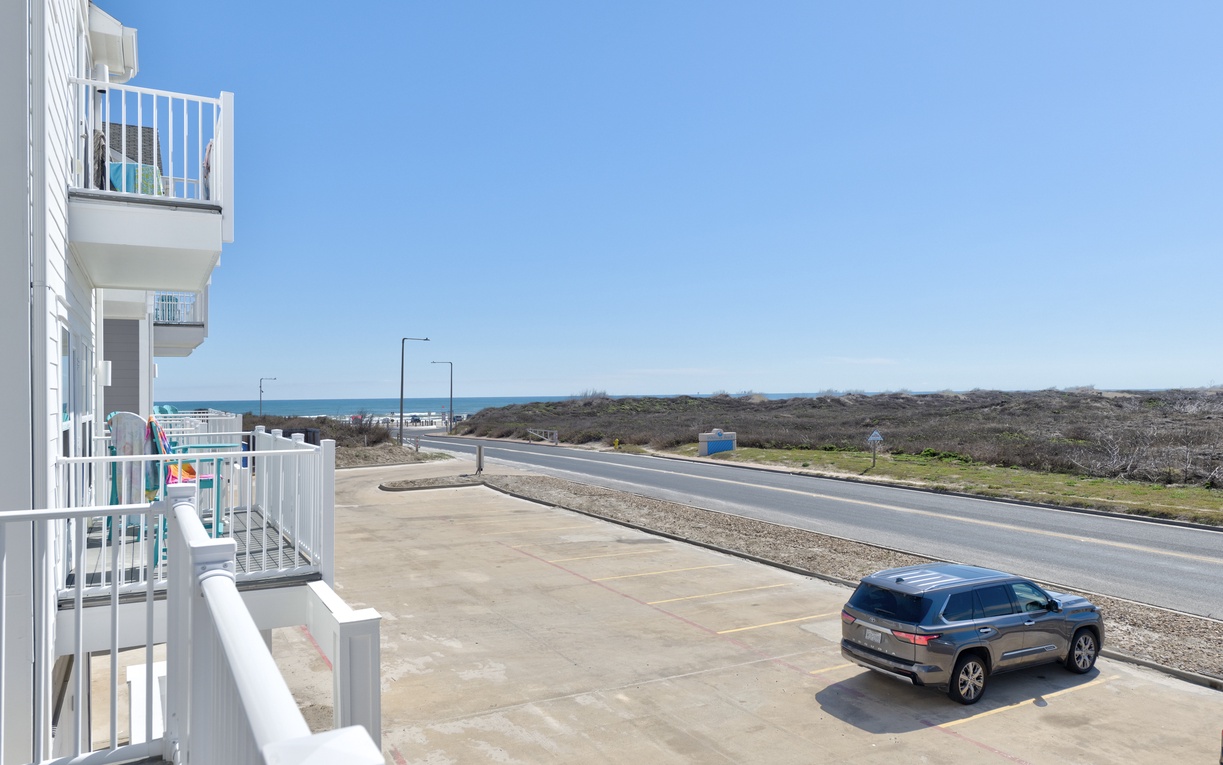White multi-story building features multiple balconies overlooking a coastal road leading toward the ocean horizon.