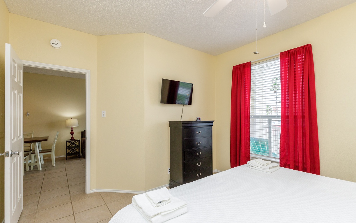 Your cozy bedroom features crisp white linens, vibrant red curtains, and a wall-mounted TV for relaxing evenings.