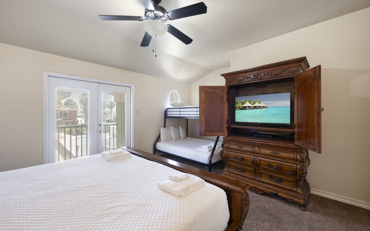 Unwind in your comfortable bedroom with plush bedding, a ceiling fan for cool breezes, and an entertainment center showcasing a flat-screen TV—plus private balcony access for fresh air and relaxation.
