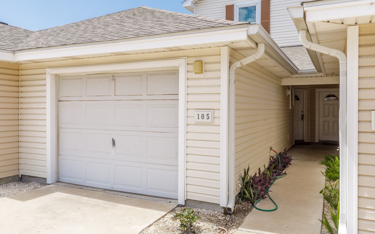 Welcoming entrance to unit 105 with covered walkway, garage parking, and charming landscaped gardens greeting your arrival.