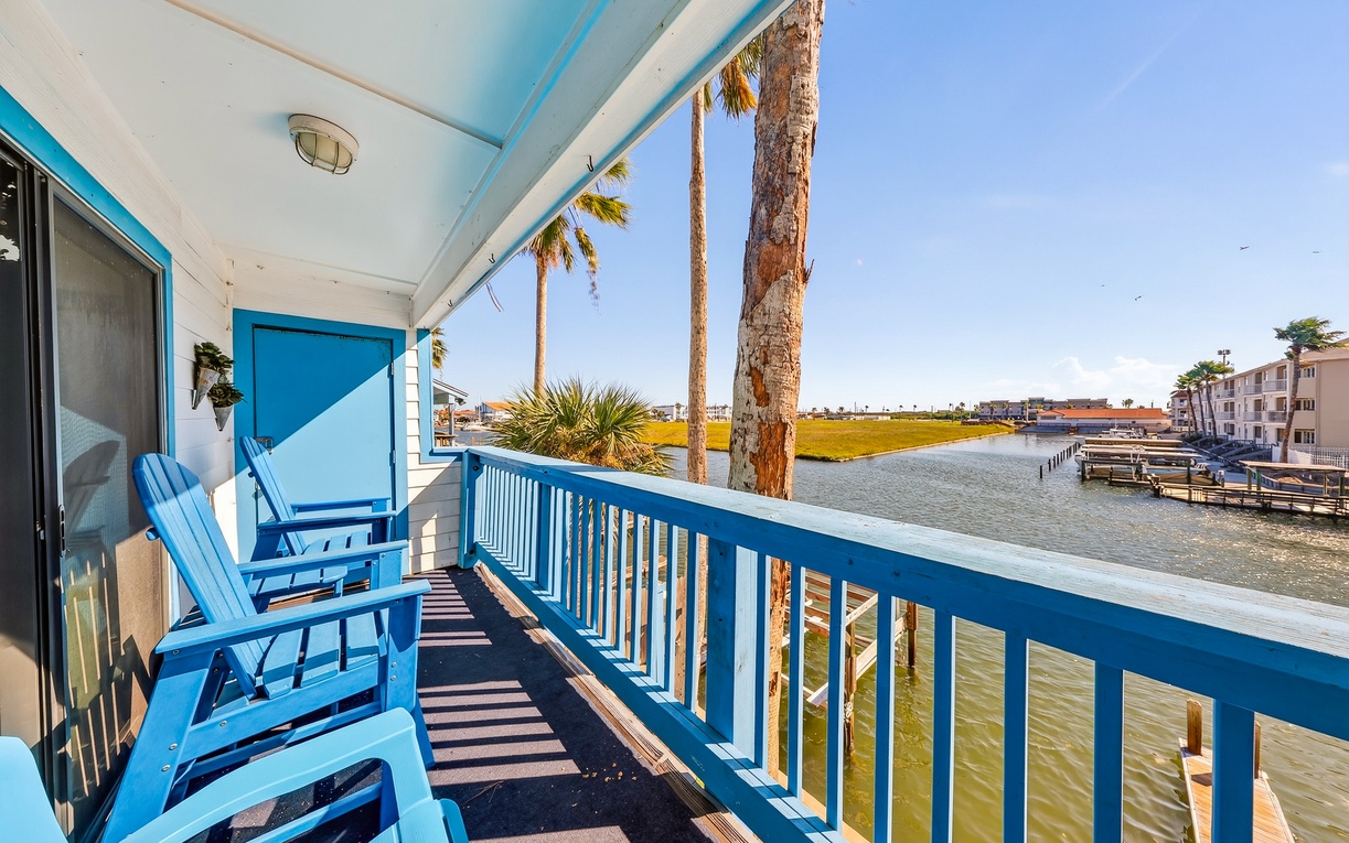 Step onto your private balcony where blue Adirondack chairs invite you to soak up the coastal sunshine and waterfront views, swaying palms framing the peaceful scene beyond.