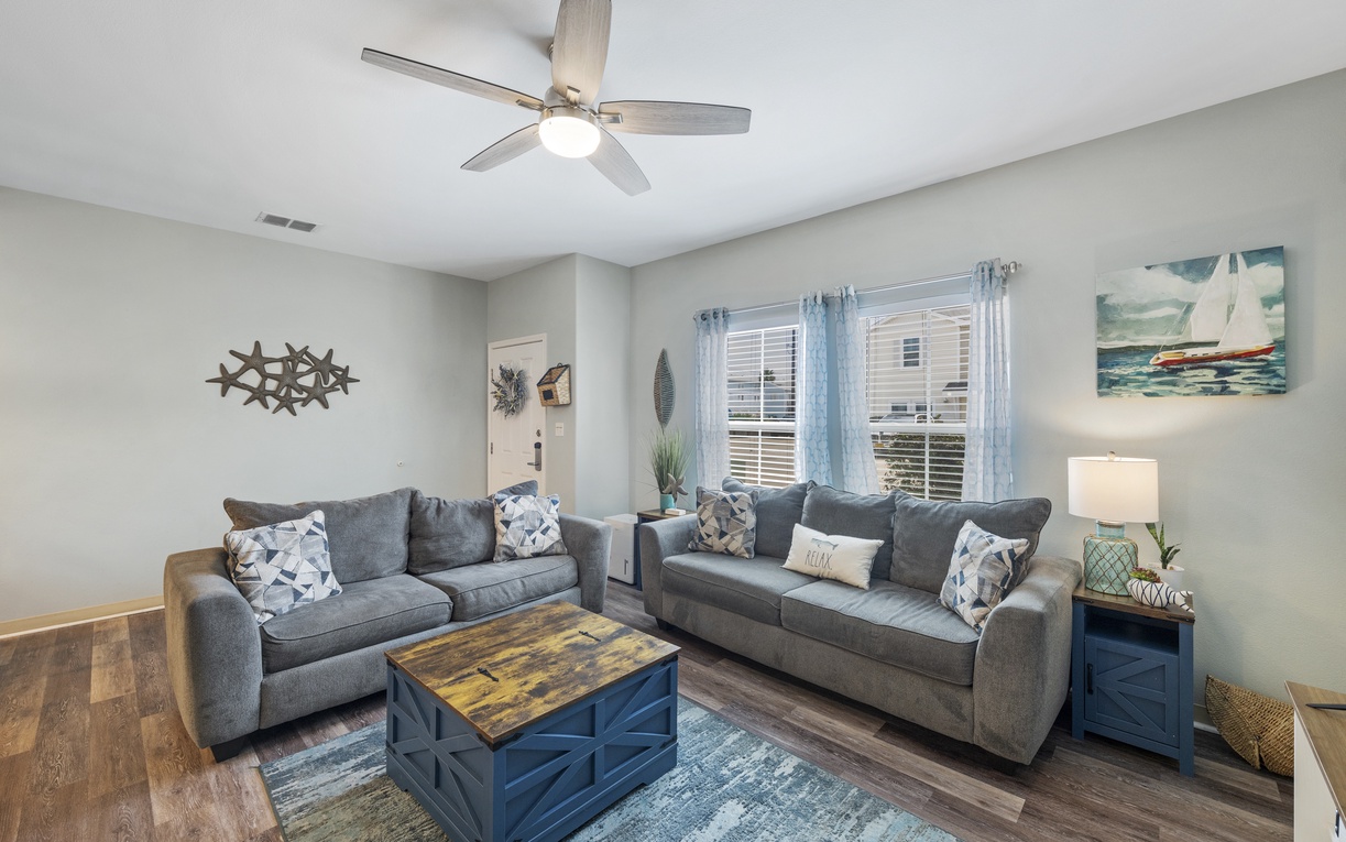 Sink into plush gray sofas under the gentle ceiling fan breeze, surrounded by charming nautical decor that makes this living space feel like home.