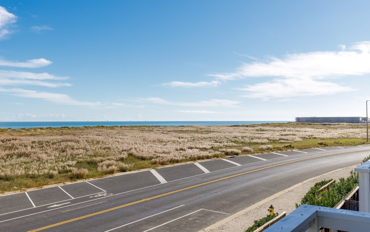 Coastal landscape stretches toward the ocean beyond quiet roadway and natural dune vegetation.
