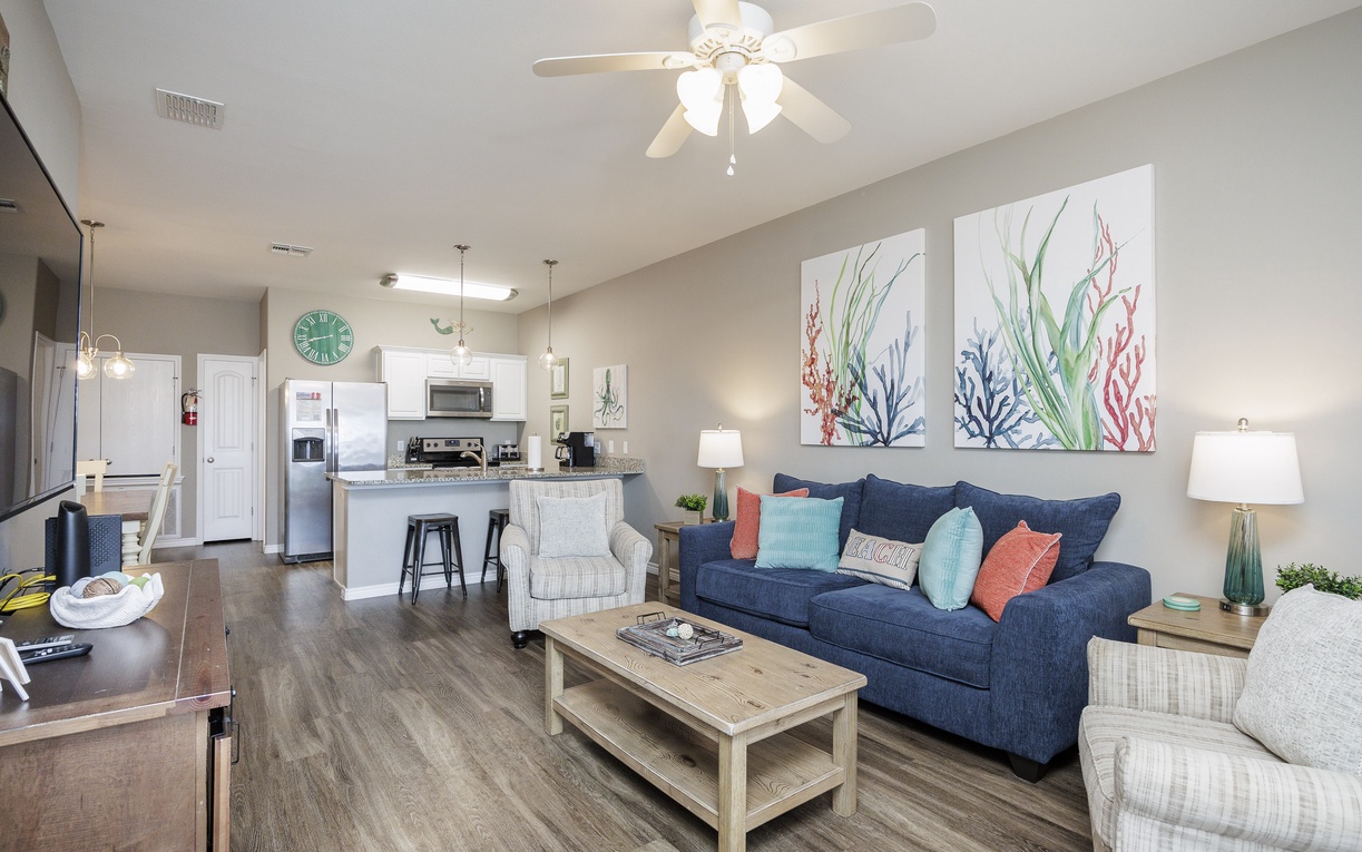 Relax in your bright, coastal-inspired living space with a plush navy sofa, charming sea-themed artwork, and an open kitchen perfect for preparing meals.