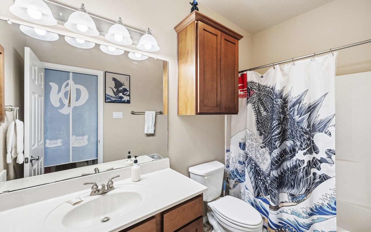 Your private bathroom features a Godzilla shower curtain, warm wood cabinetry, and fresh white linens waiting to make you feel at home.