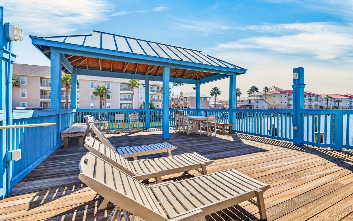 A spacious wooden deck with a vibrant blue pergola awaits your arrival, offering sunbeds for lounging and dining areas perfect for gathering. Palm trees and nearby property buildings frame the open coastal atmosphere.