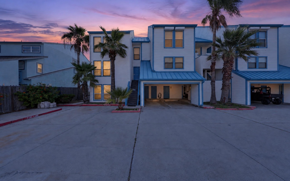 A modern multi-story vacation home framed by palm trees at twilight, with warm lights glowing through the windows and convenient parking right at the entrance.