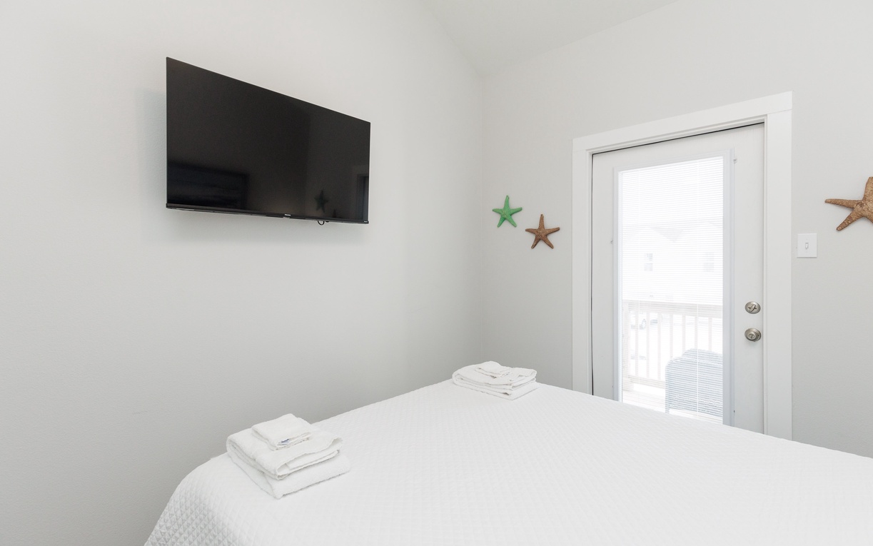 Unwind in your serene bedroom with coastal charm, featuring a wall-mounted TV, queen size bed, and fresh linens and towels for your comfort.