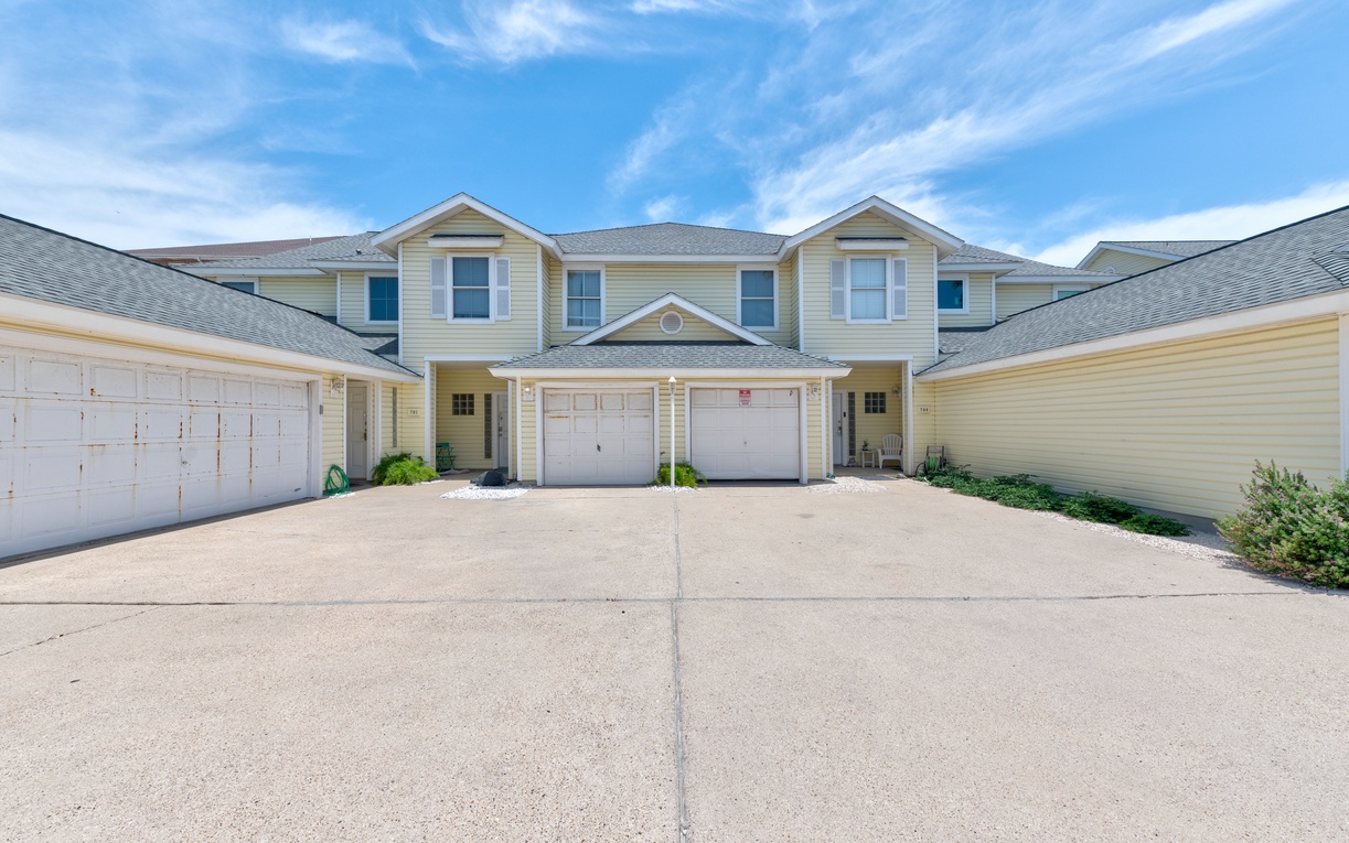 Bright coastal townhome with convenient garage parking and charming dormer windows under expansive blue skies.