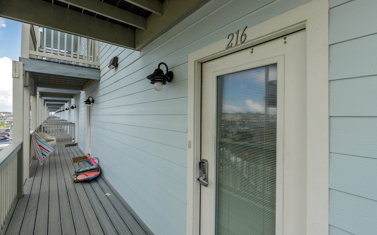 Unit 216 entrance with private balcony overlooking the peaceful surroundings. Modern light blue siding creates a welcoming coastal atmosphere for your getaway.