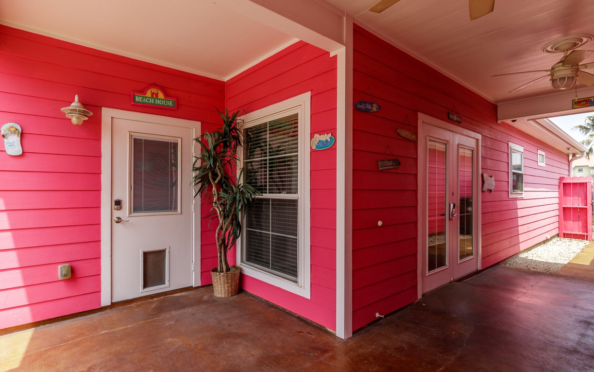 Bright coral-colored beach house exterior with charming covered porch and tropical plant welcomes you to your coastal getaway.