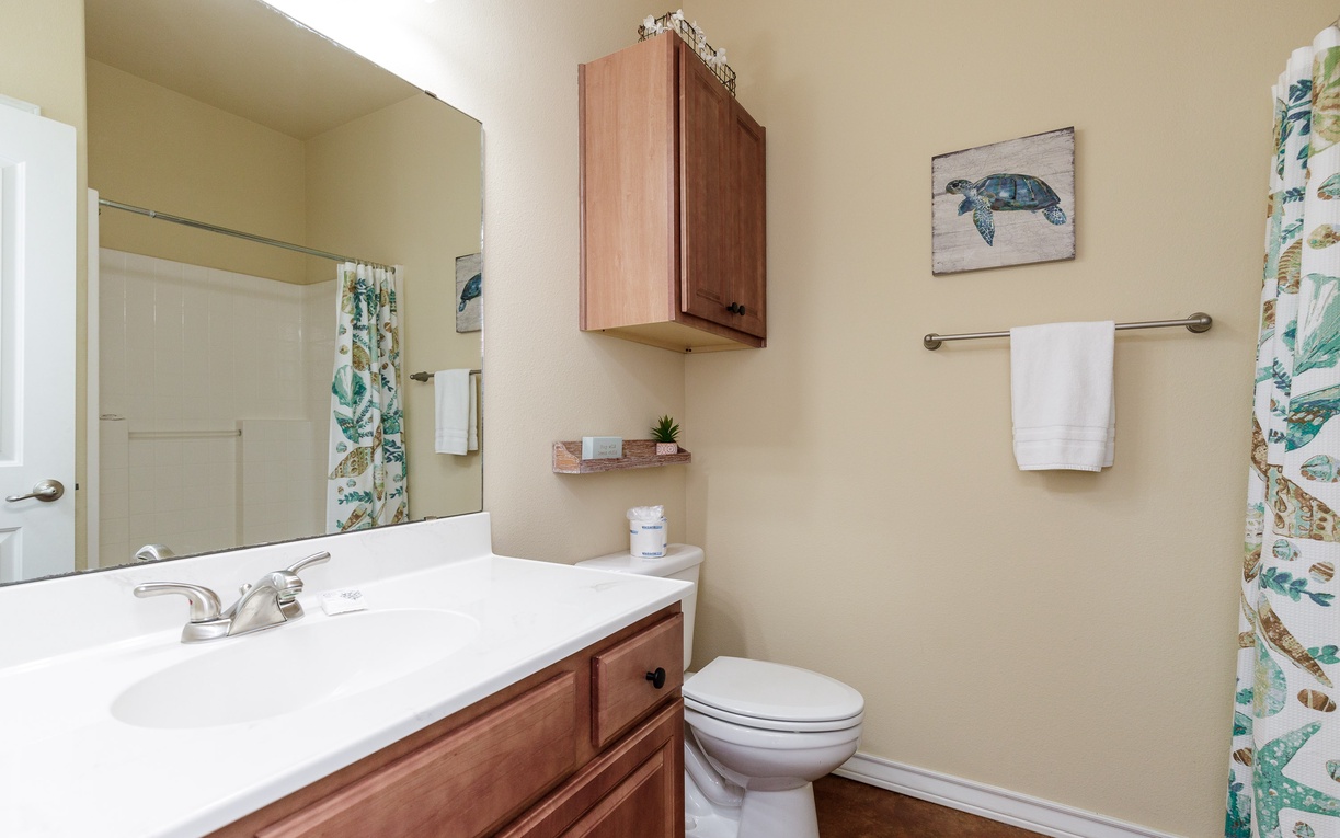 Your private bathroom features warm wood cabinetry, coastal turtle artwork, and tropical shower curtains that set a beachy, relaxing tone.