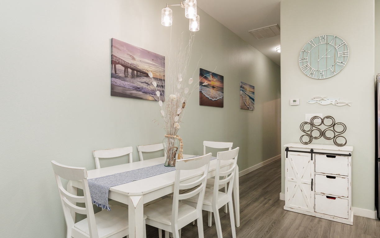 Gather around your elegant white dining table, where coastal artwork and warm pendant lighting create the perfect setting for memorable meals together.
