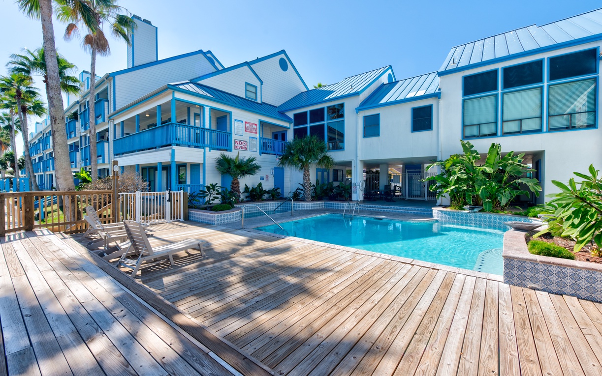Tropical resort complex featuring a beautiful swimming pool surrounded by wooden deck and lush palm trees under clear blue skies.