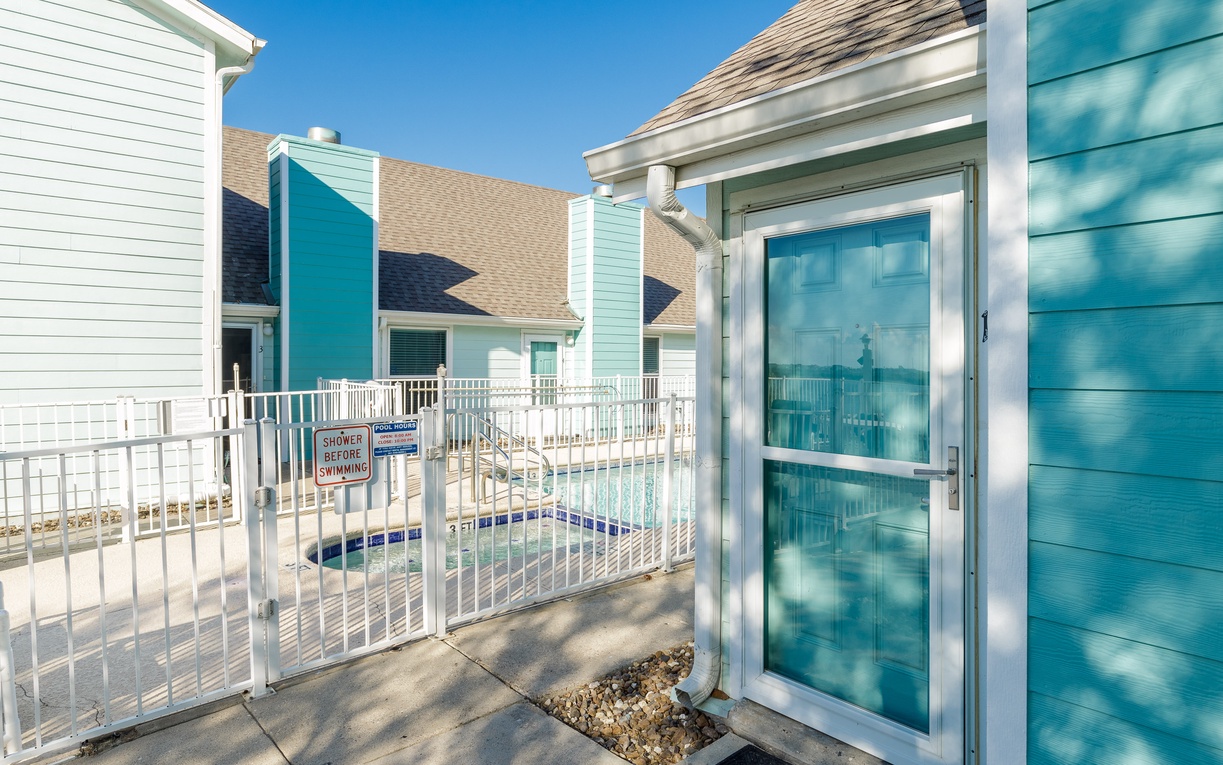 Bright turquoise building exterior with glass doors leading to a fenced pool area under clear blue skies.