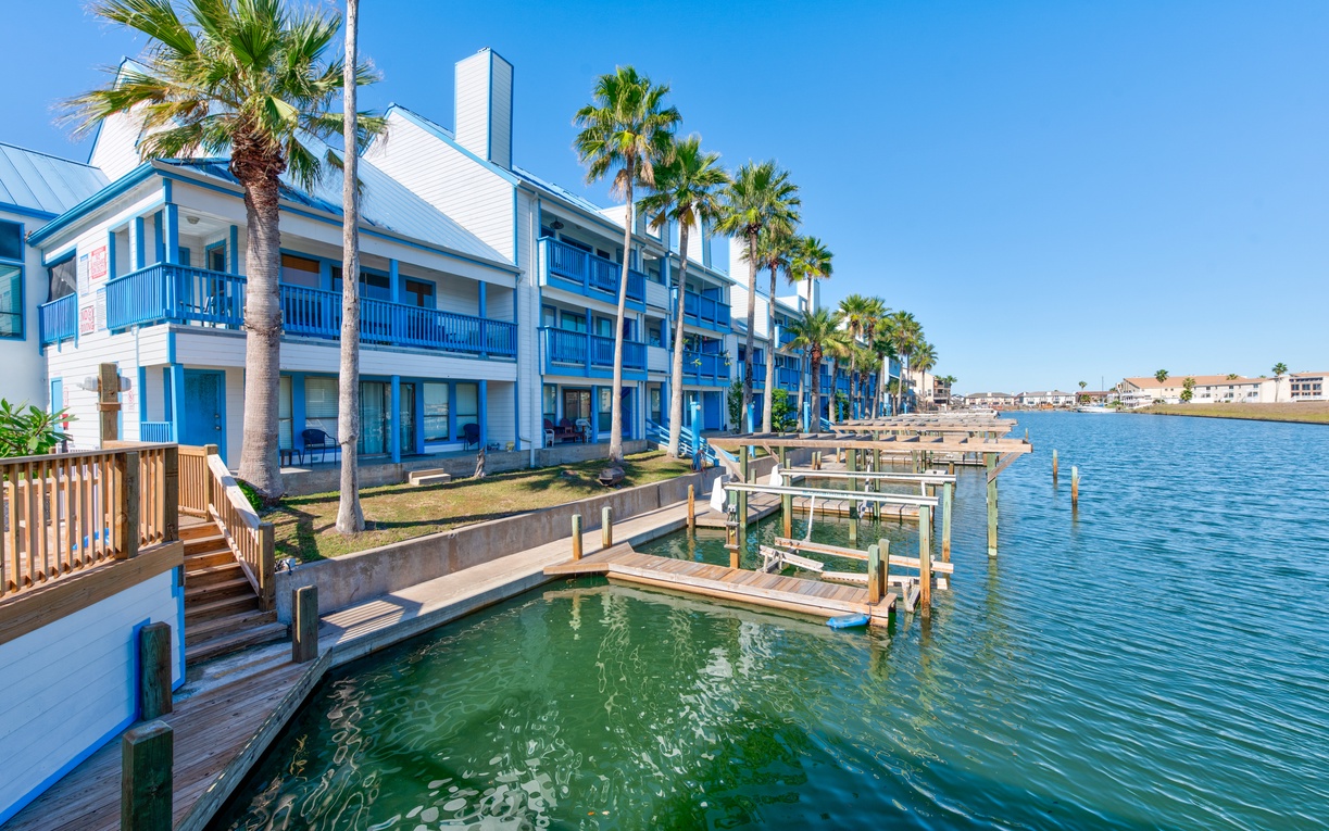 Waterfront property featuring palm-lined buildings with boat docks and direct water access under bright blue skies.