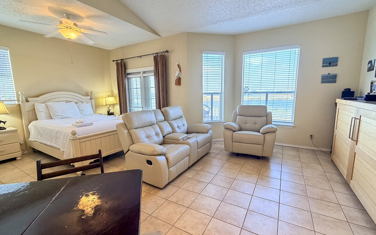 Relax in your spacious suite with plush reclining chairs, a comfortable bed, and warm natural light streaming through multiple windows.
