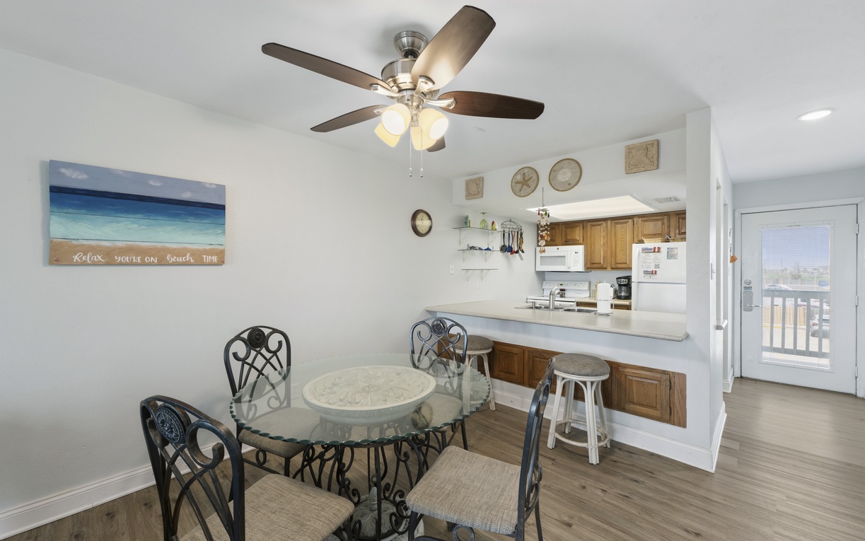 Gather around this cozy dining area, where coastal vibes and beach time decor set the tone for your getaway, complete with an open kitchen and balcony access nearby.