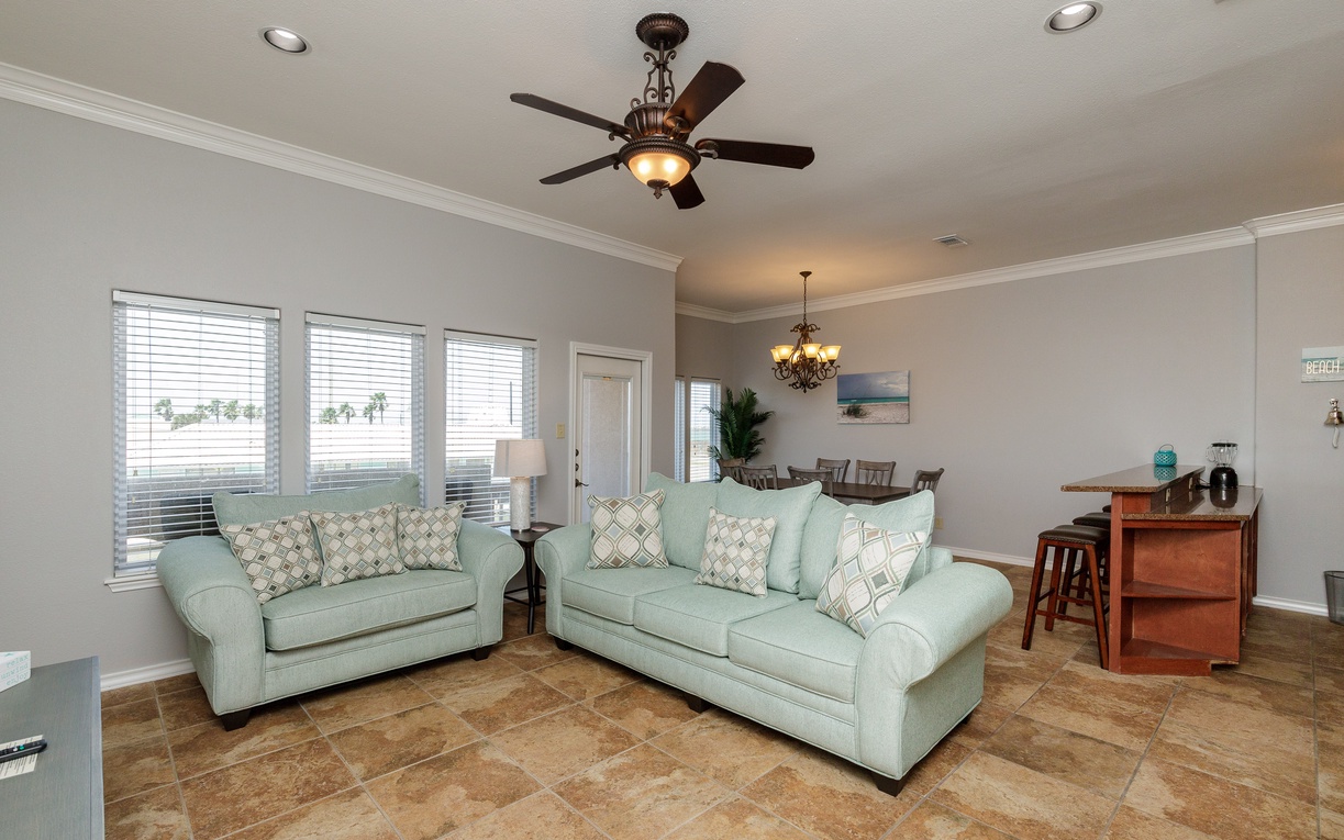 Relax in your spacious living room with plush mint sofas, elegant ceiling fan, and bright windows overlooking palm trees.