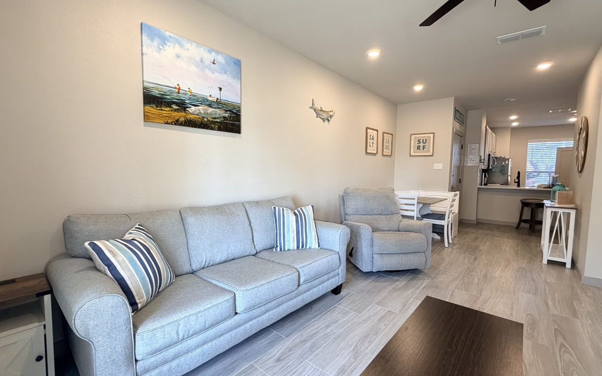 Your spacious living room invites relaxation with its comfortable grey sofa, coastal artwork, and open-concept flow into the kitchen.