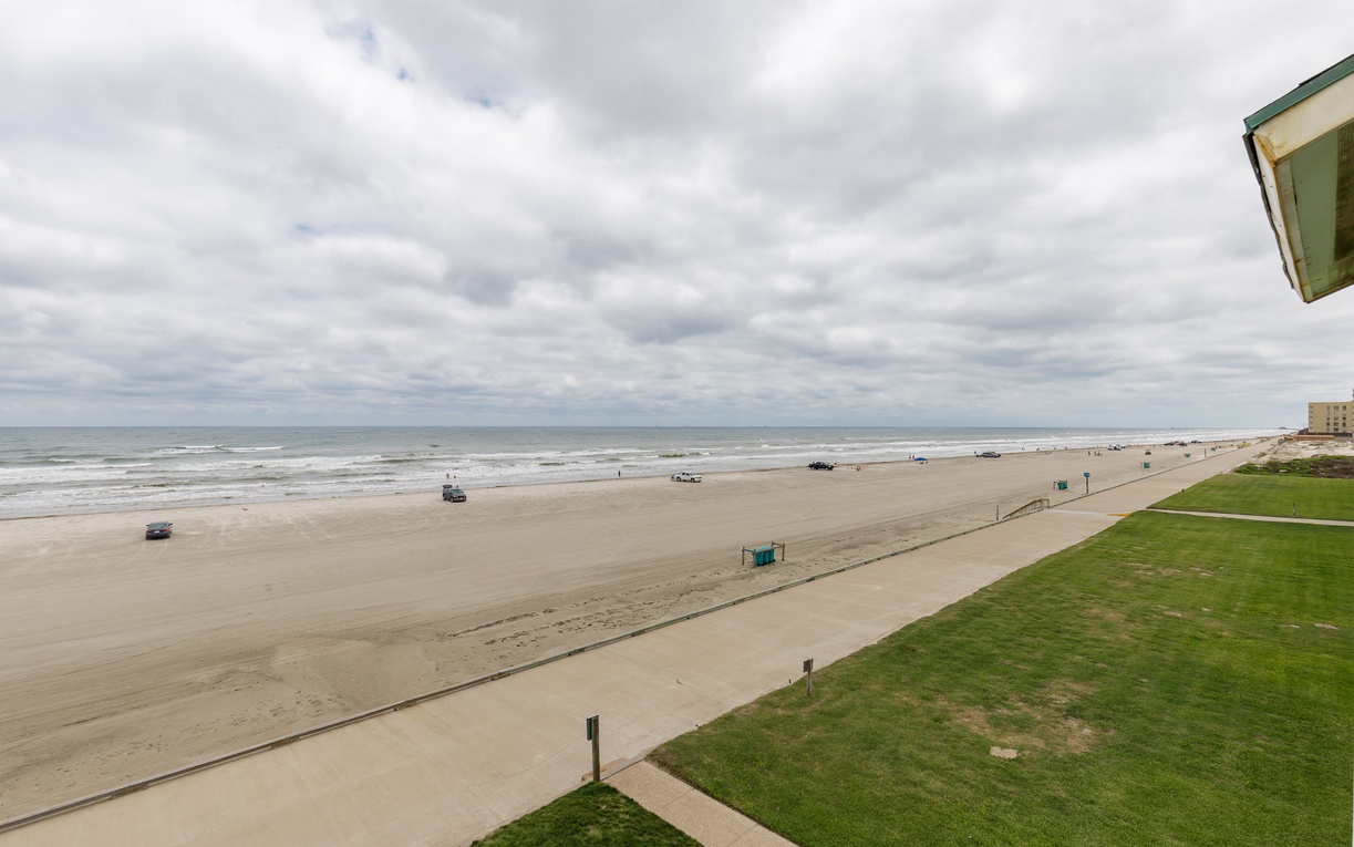 Wide sandy beach stretches endlessly along the coastline, with gentle waves and scattered beachgoers under an expansive cloudy sky.