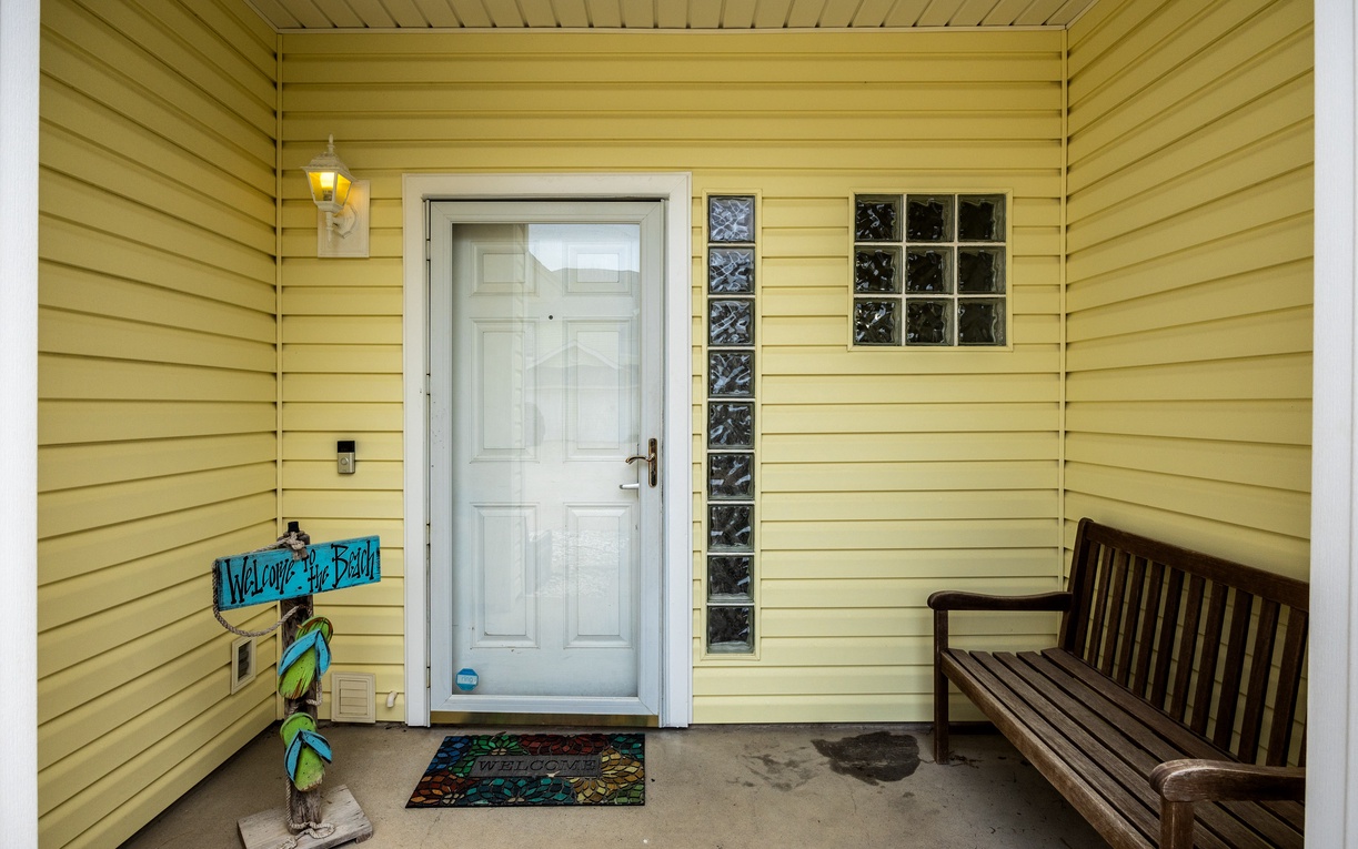 Step onto your covered porch where a welcoming bench and cheerful turquoise sign create the perfect first impression of your beach getaway.