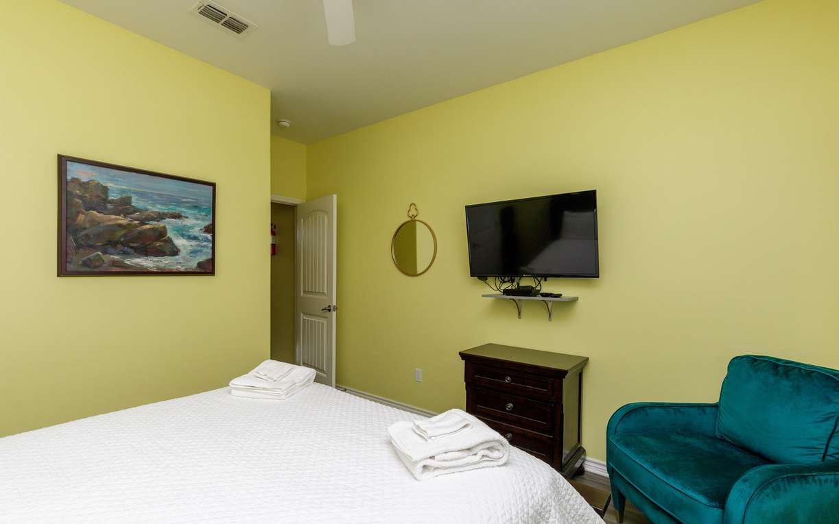 Unwind in your cheerful bedroom featuring a plush teal armchair, wall-mounted TV, and crisp linens for restful nights.