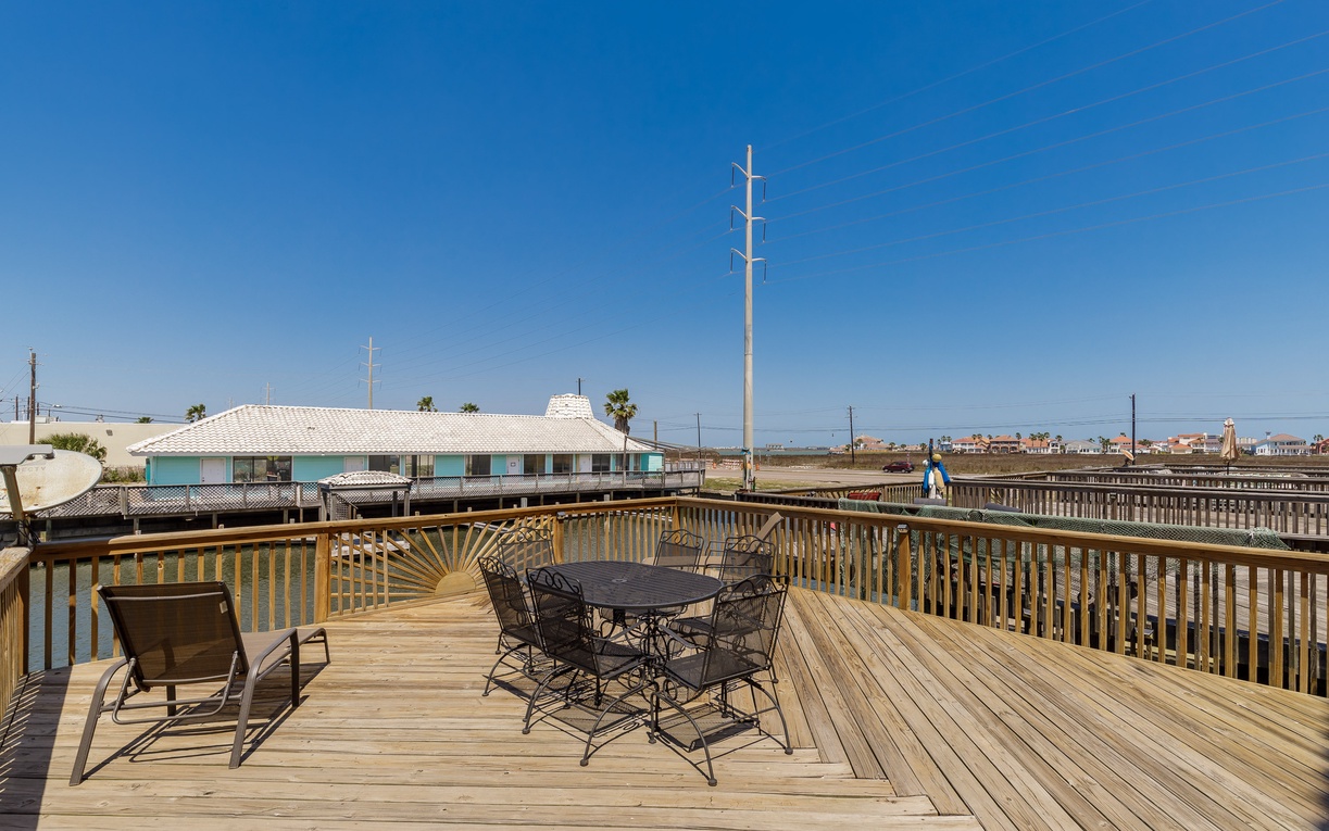 Spacious wooden deck with dining table and lounge chairs overlooking coastal neighborhood under clear blue skies.