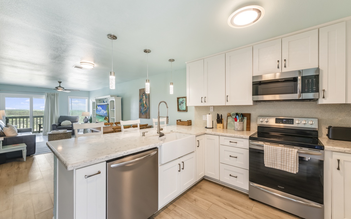 Your fully-equipped kitchen features bright white cabinetry, sleek stainless appliances, and modern pendant lighting—perfect for preparing meals while enjoying breezy coastal views from the open living area.