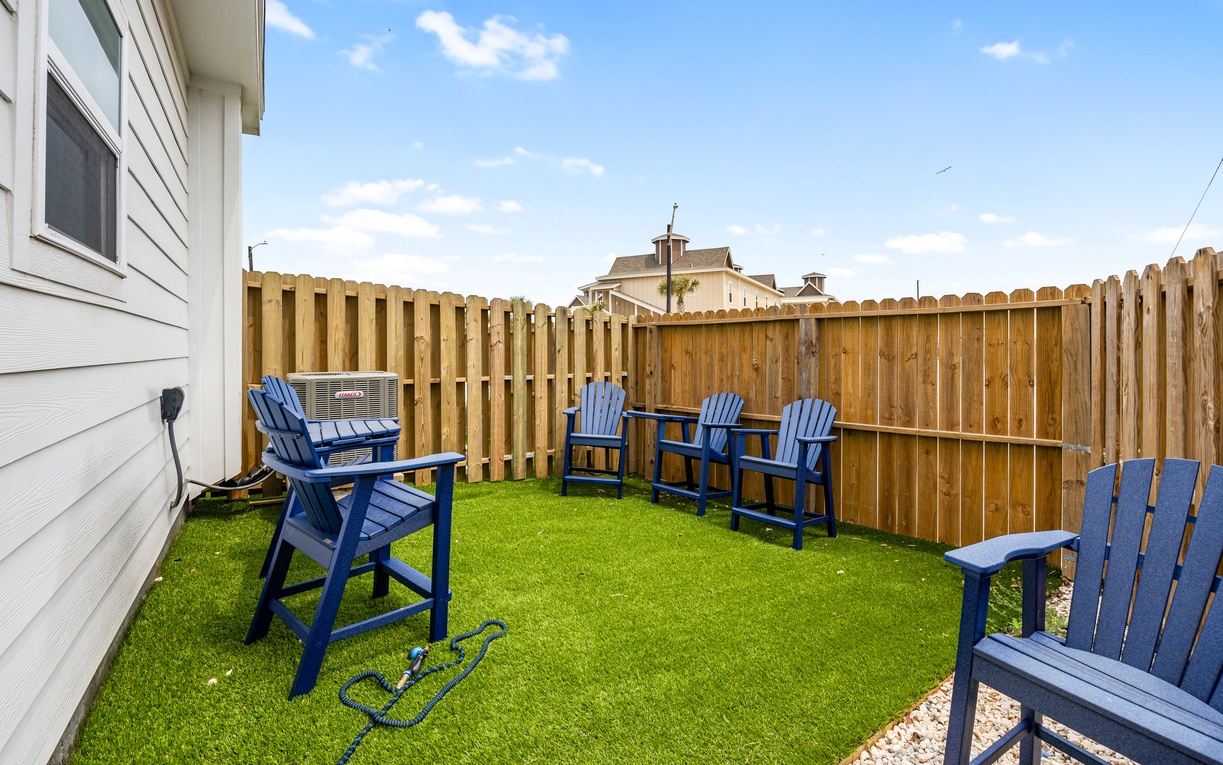 Your private backyard retreat features vibrant blue Adirondack chairs on lush artificial turf, perfect for morning coffee or evening relaxation in your own peaceful space.