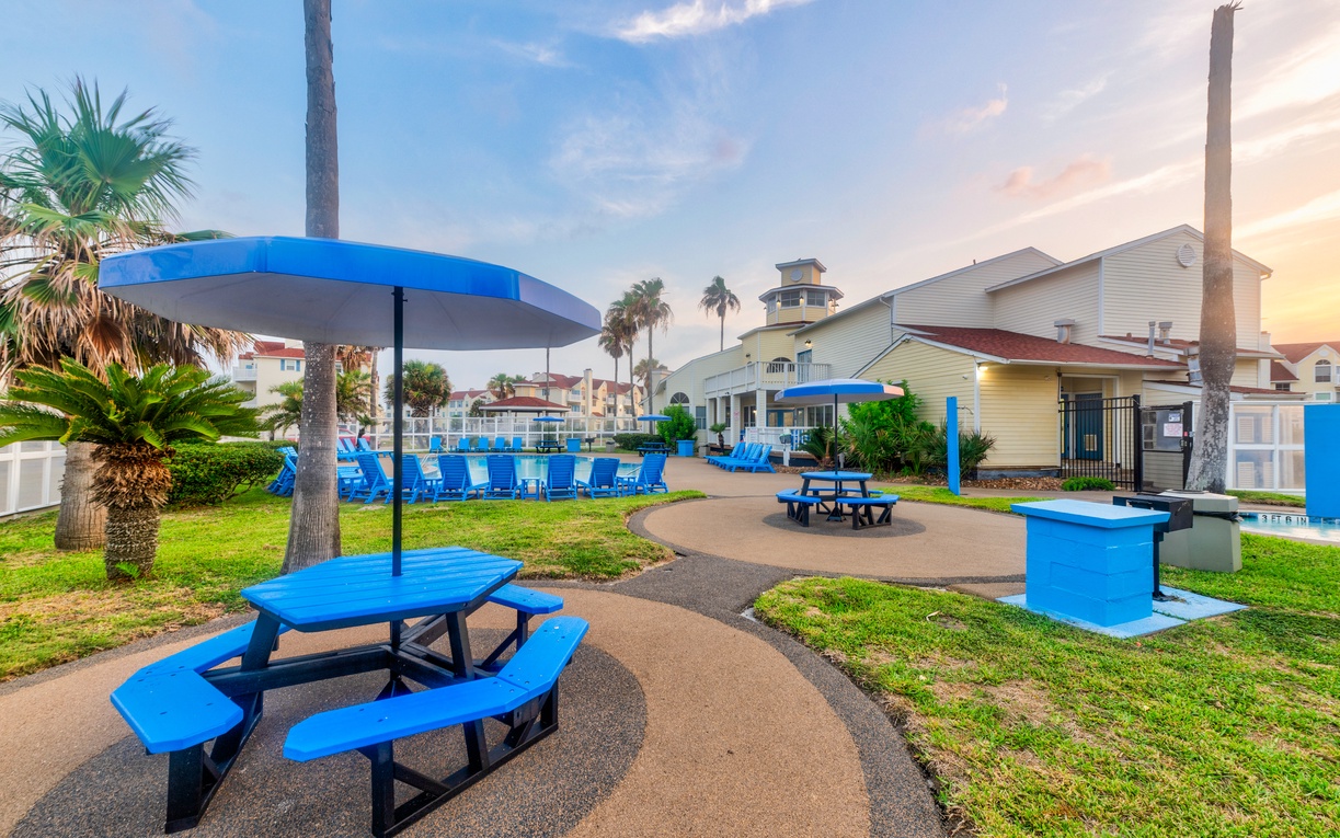Tropical resort amenities feature blue picnic tables, BBQ facilities, and palm-lined grounds with colorful buildings nearby.