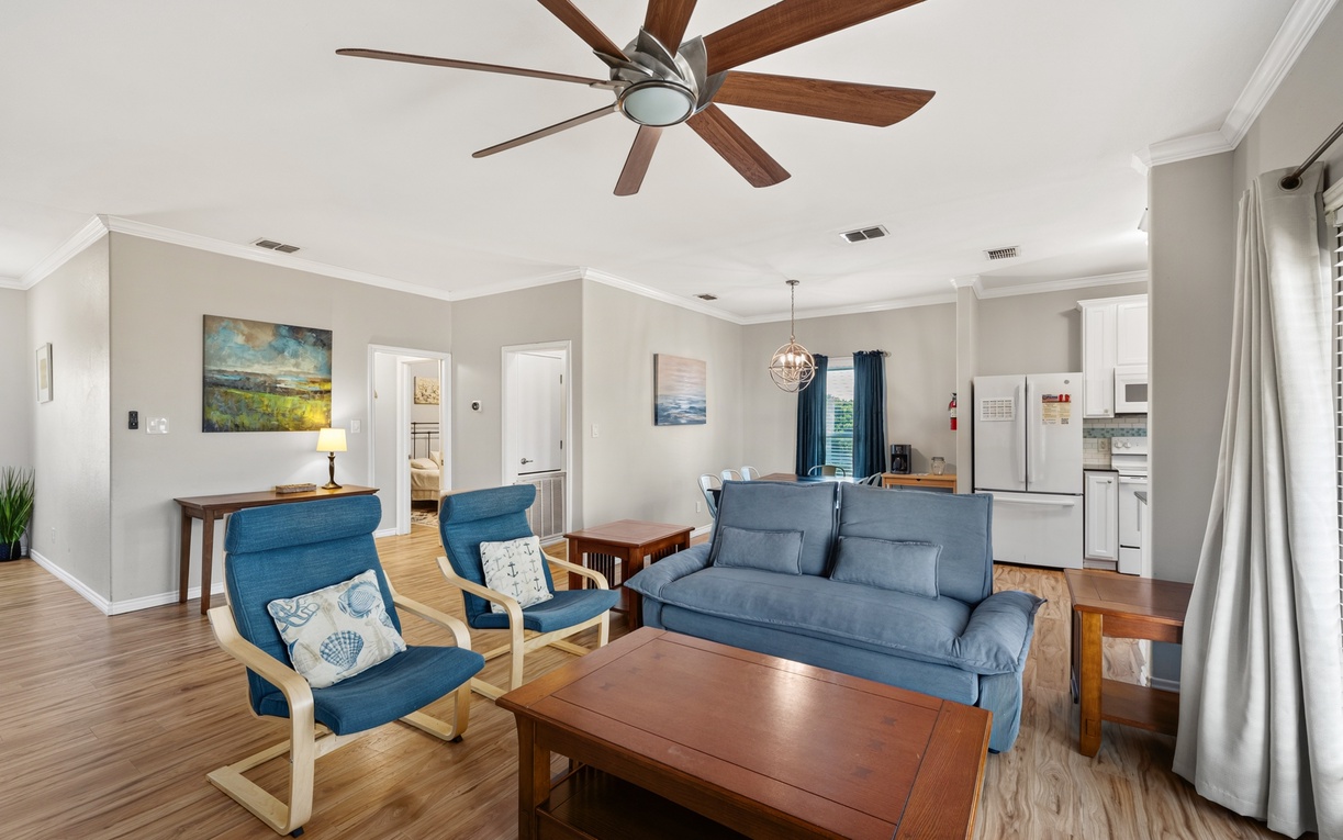 Step into your bright, open living space where coastal blues meet warm wood tones. Relax under the sweeping ceiling fan, sink into the plush sofa, and enjoy the seamless flow into the kitchen—your perfect home base for coastal escapes.