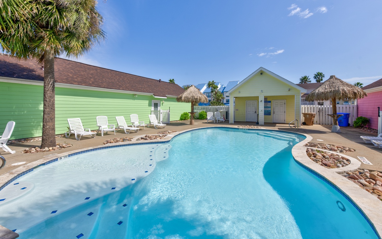 Colorful beach cottages surround a sparkling swimming pool with tropical palm trees and clear blue skies overhead.