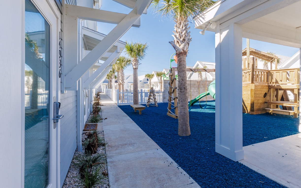 Modern coastal property featuring playground area with palm trees and covered walkways under bright blue skies.