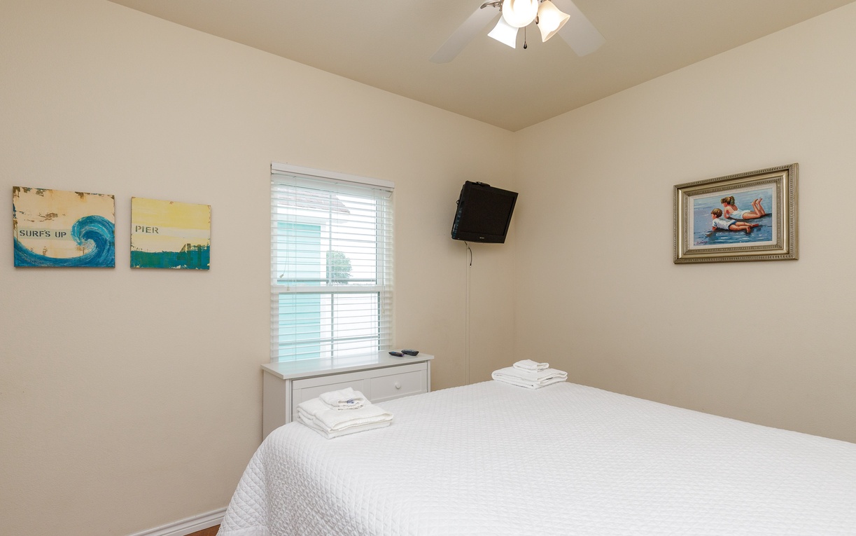 Relax in your peaceful bedroom featuring a comfortable bed, wall-mounted TV, and charming coastal artwork that sets the perfect vacation mood.