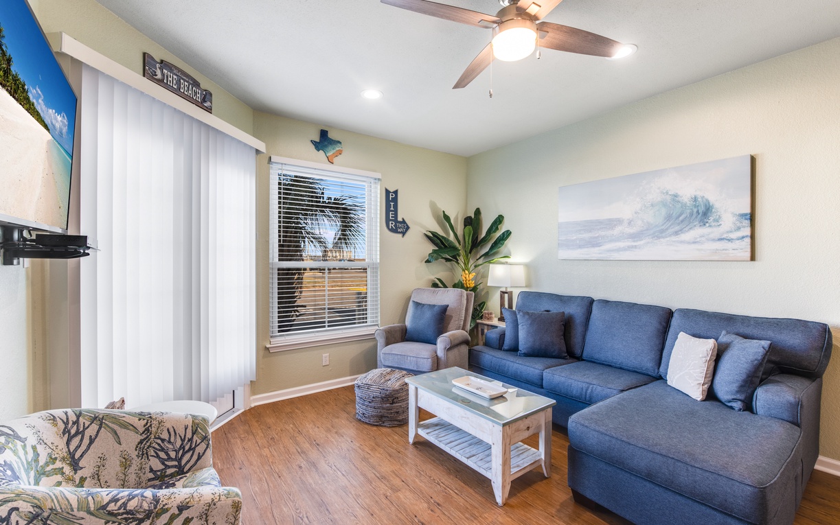 Relax in your coastal-themed living room with comfortable blue sectional seating, tropical artwork, and ceiling fan for perfect comfort.